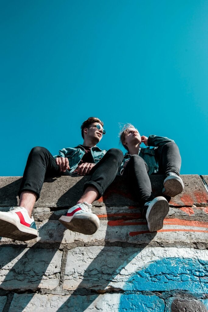 Two young people sitting on a stone wall under a vibrant blue sky, enjoying a sunny day together.